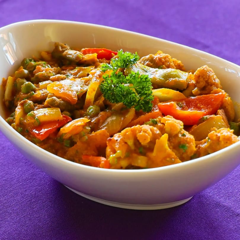 A white bowl filled with a vibrant Indian vegetable curry, garnished with fresh parsley, on a purple tablecloth.