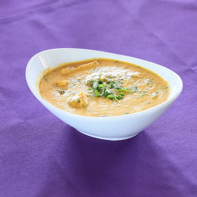 Zaika Indian Cuisine's creamy Indian curry, garnished with fresh herbs, in a white bowl on a purple tablecloth.