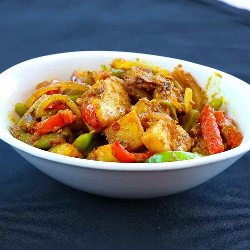 A white bowl filled with Zaika Indian Cuisine's Aloo Capsicum, featuring potatoes, red and green bell peppers.